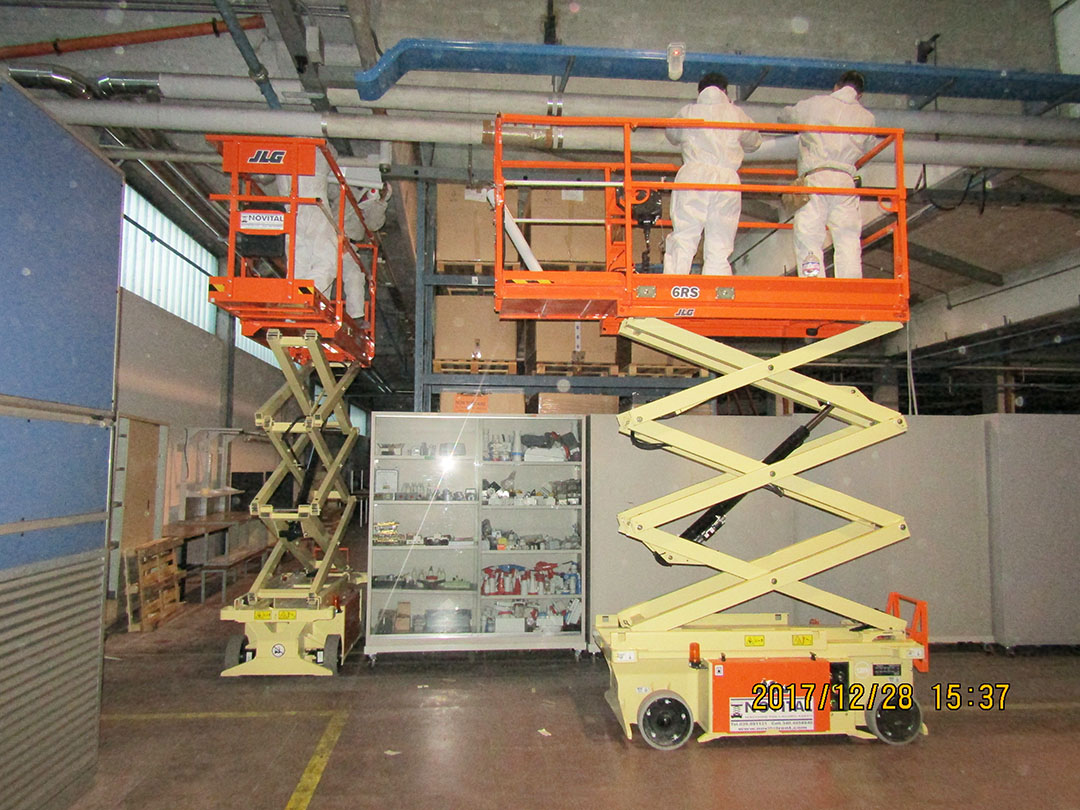 Operai all'opera sopra piattaforme elettriche con estensione a pantografo per la messa in sicurezza di tubazioni aeree coibentate in cemento amianto compatto  ubicate all'interno di una fabbrica.
